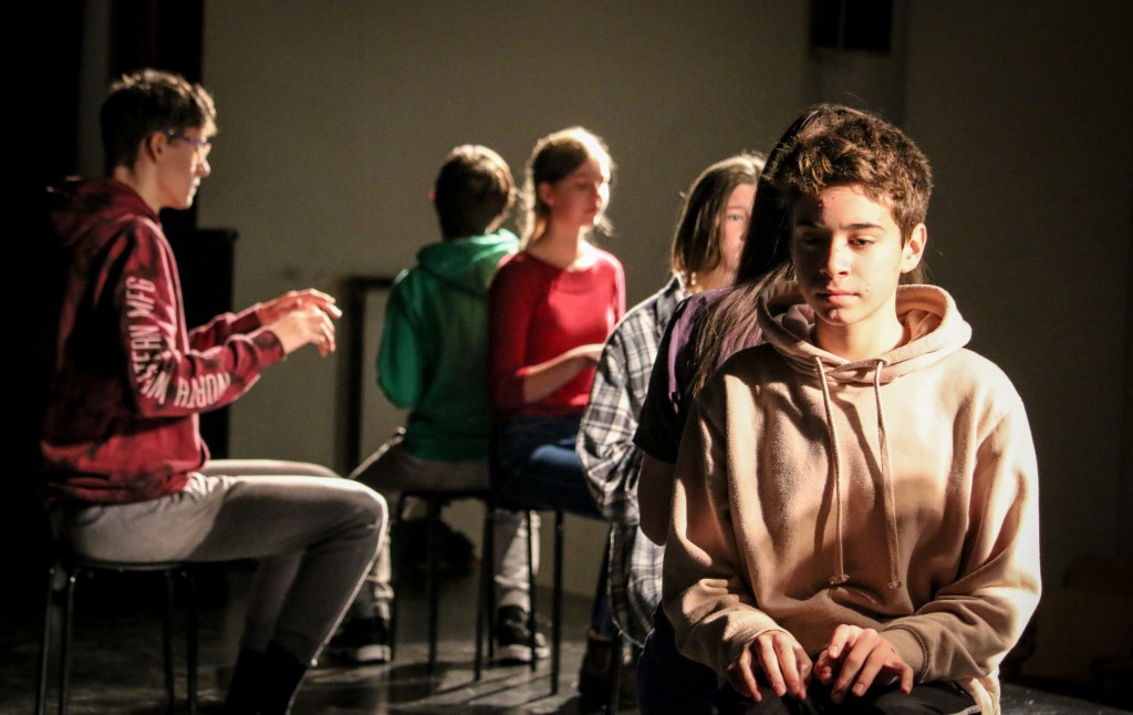 6 youth sit and type on imaginary keyboards. They sit back to back and face in different directions.