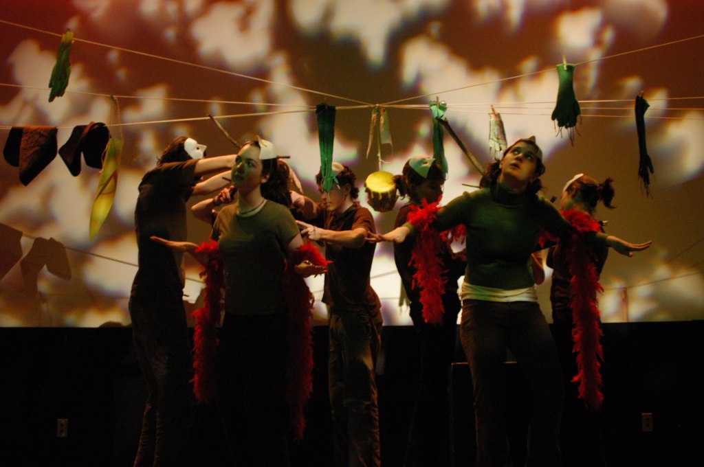 8 youth stand in a ciircle, back to back. They wear green with red scarves. They have masks on their heads. There are strings with objects dangling above them,