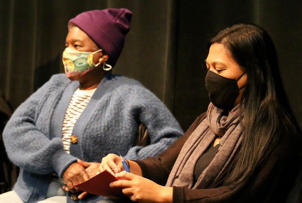 Jo-Anne wears a blue sweater and purple cap and has her hands clasped; beside her, Carmelle makes notes; she wears a brown sweater with a tan scarf. They wear masks