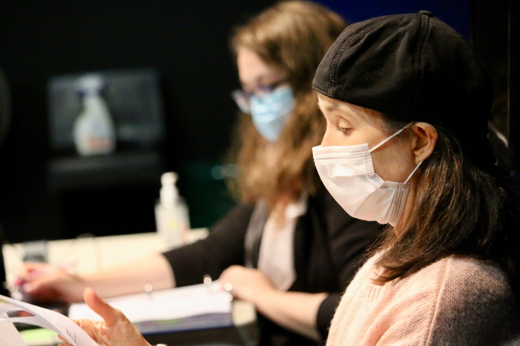 two women read scripts in a rehearsal studio; each wear masks and sit at a white table
