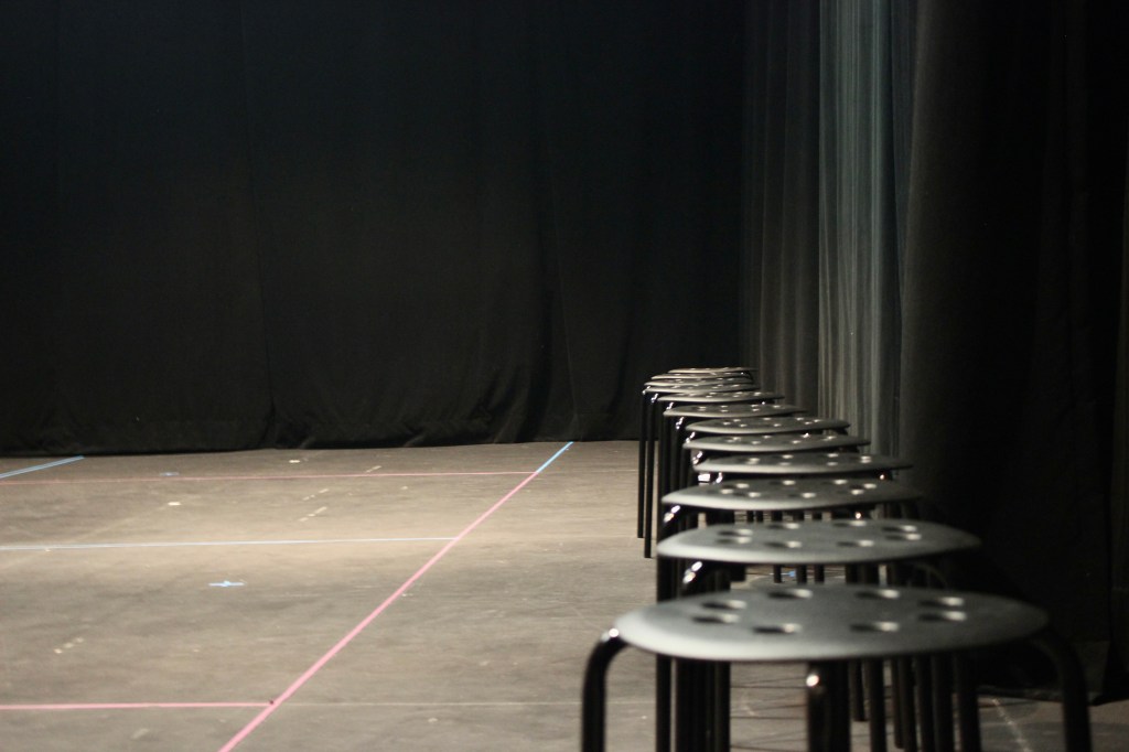 12 black stools with holes in their surface and 4 legs sit in a row along a black curtain. The room is also black. The floor has coloured tape marks on its surface