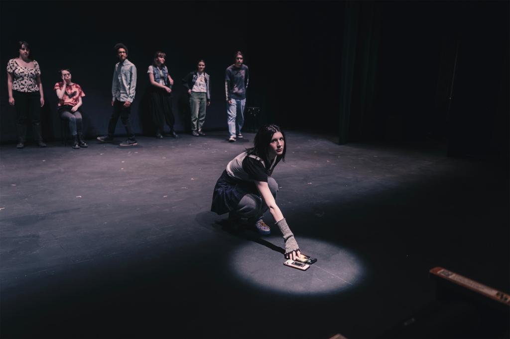 [Image description: a youth puts their cell phone into a spotlight while looking out at the audience. Other youth are upstage behind them and watch.]