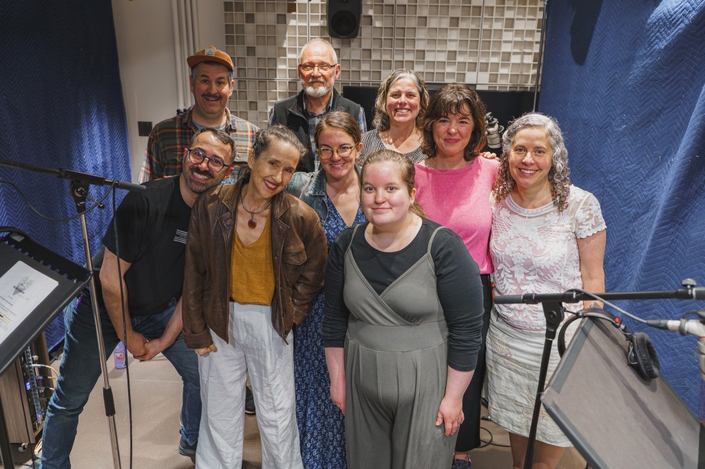 9 people stand in a recording studio and smile at the camera, with a background with blue curtains and on each side, microphones.