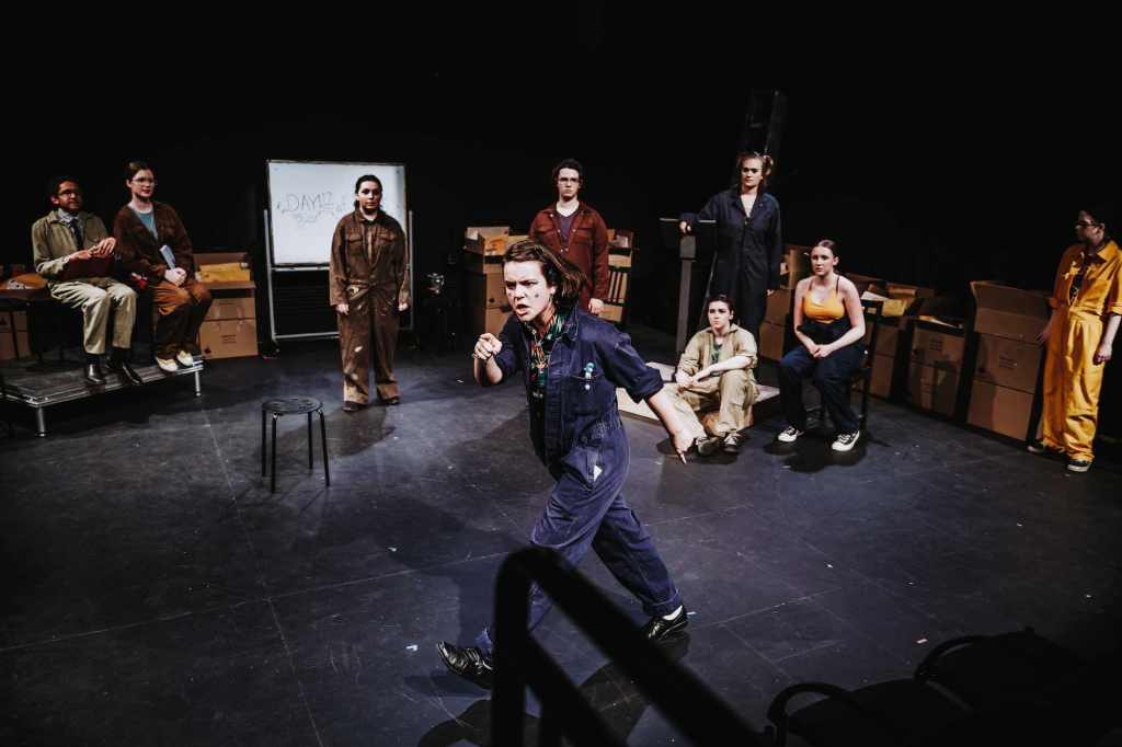9 actors wearing different colored coveralls perform on a stage. One actor walks across the stage looking determined while the others watch and react.