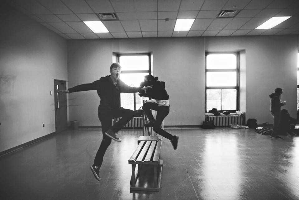 Black and white photo of two youth jumping over a bench. Two windows in the background. A classroom studio.