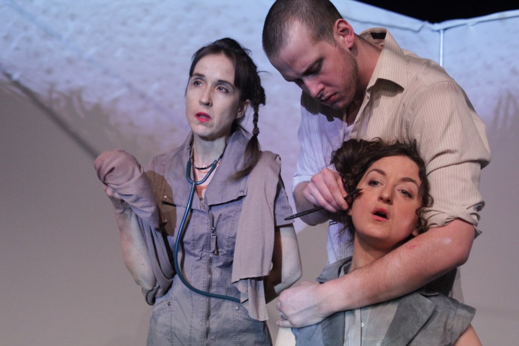 Three characters in a play. One holds another in a neck grip, the other, wearing a stethoscope, looks out. Colors of clothing are muted greys and browns.
