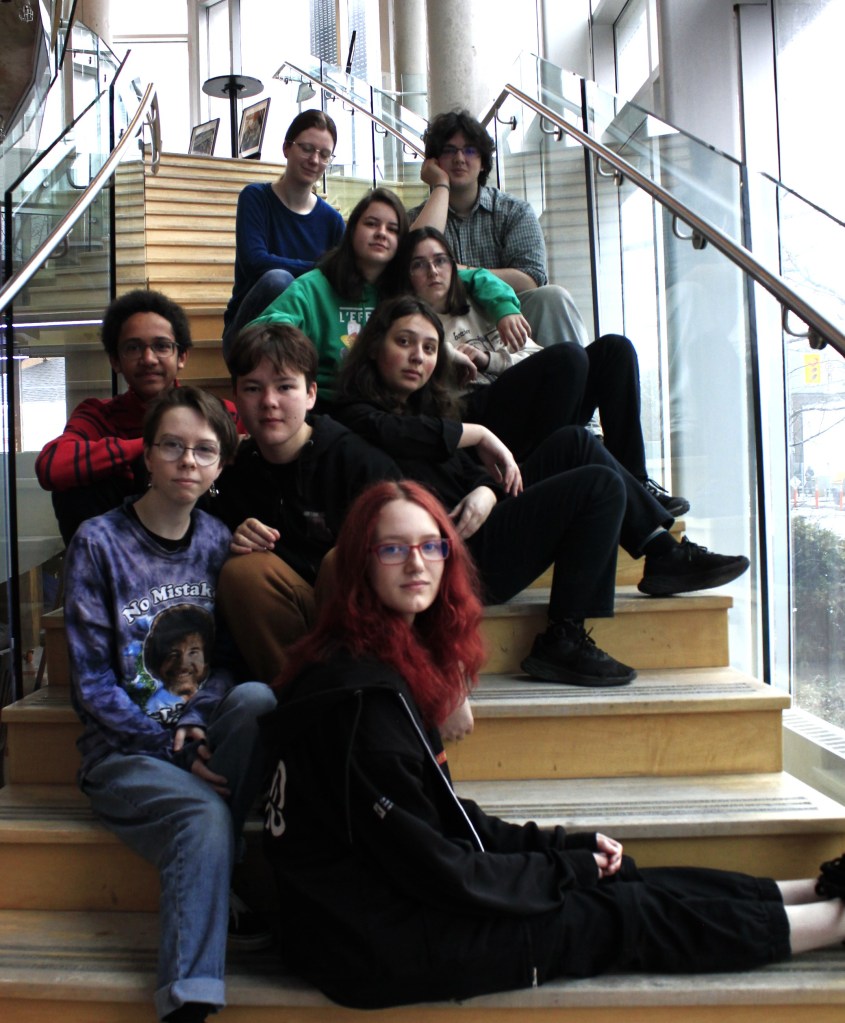 A group of 10 youth sit on beige coloured stairs and look at the camera. They are dressed in casual clothing. There are glass windows on one side.