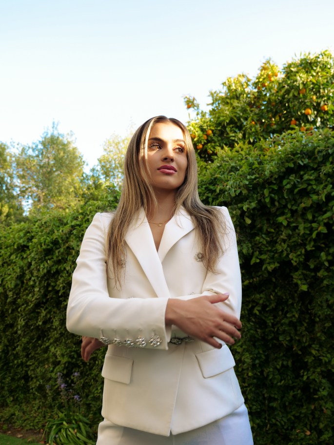 [Image Description: Youth with long brown hair wearing a white blazer stands outside in front of a hedge.]