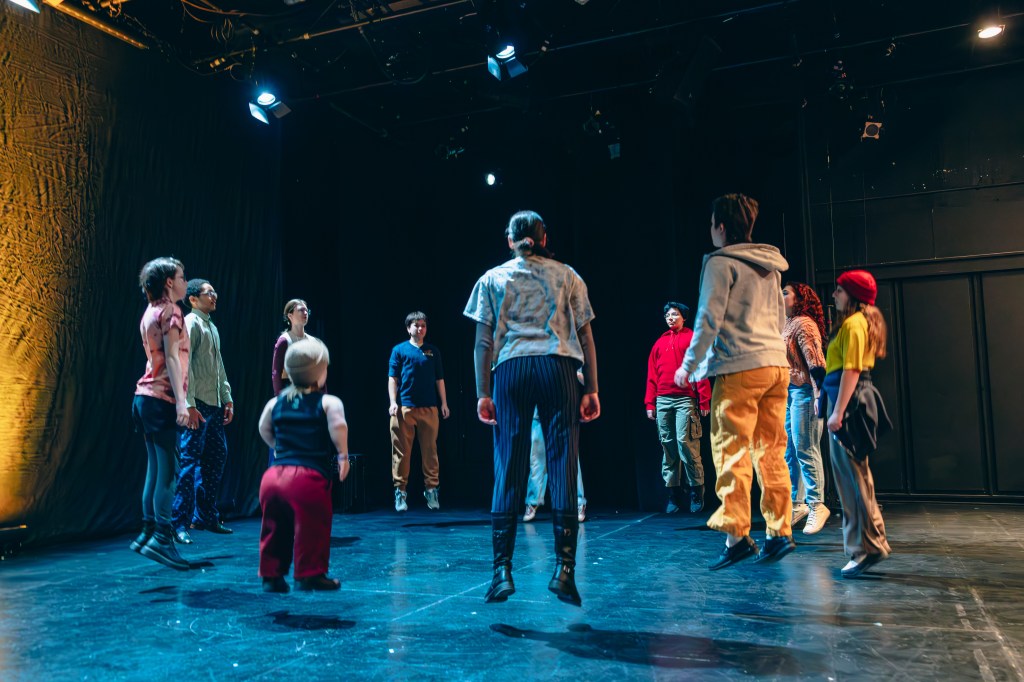11 youth jump in a circle. They are on a stage and wear different coloured clothing