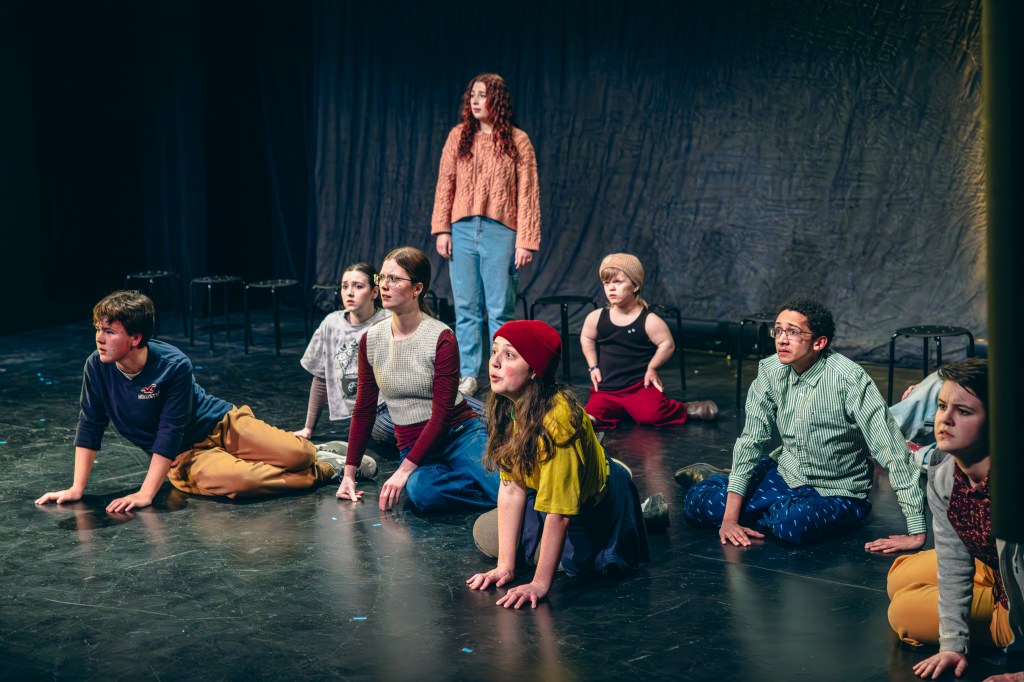 9 youth sit and one stands on a stage and look out at the audience in hope/plea.
